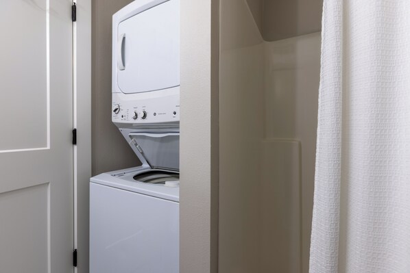 A compact, white, front-loading washer and dryer unit in a small laundry room with a plain door and a white curtain.