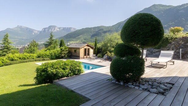 Una terraza de madera con piscina, sillas de descanso y un edificio pequeño, rodeado de vegetación y montañas.