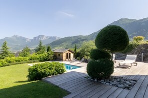 Eine Holzterrasse mit Swimmingpool, Liegestühlen und einem kleinen Gebäude, umgeben von viel Grün und Bergen.