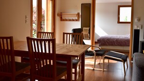 Una habitación con una mesa de madera, sillas, un banco, una cama con cabecera, un espejo y una ventana con vista a árboles.
