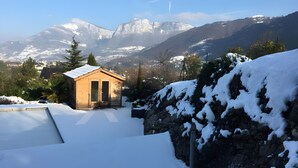 Una cabaña de madera con techo cubierto de nieve, una puerta metálica y un ventanal pequeño. Al fondo, montañas nevadas y un cielo despejado.