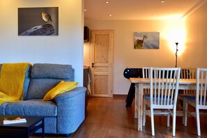 Een woonkamer met een bank, een eettafel met stoelen, een houten deur en een aan de muur hangend schilderij.