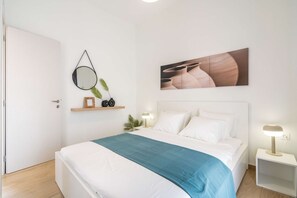 Une chambre à coucher bien rangée, avec un lit, des tables de chevet, un miroir fixé au mur, un tableau encadré et une petite étagère ornée d’objets décoratifs.