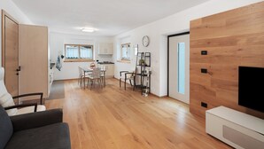 A modern living room with a sofa, a TV, wooden flooring, a dining area with chairs, and a kitchen in the background. - Apartment 'Appartamento Rododendro' with Mountain View, Private Terrace and Wi-Fi (San Tomaso Agordino)