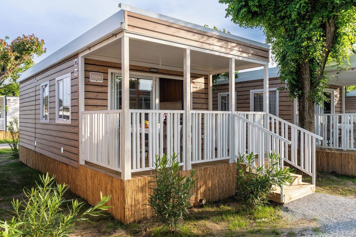 A mobile home with a porch, wooden exterior, and a fenced yard.
