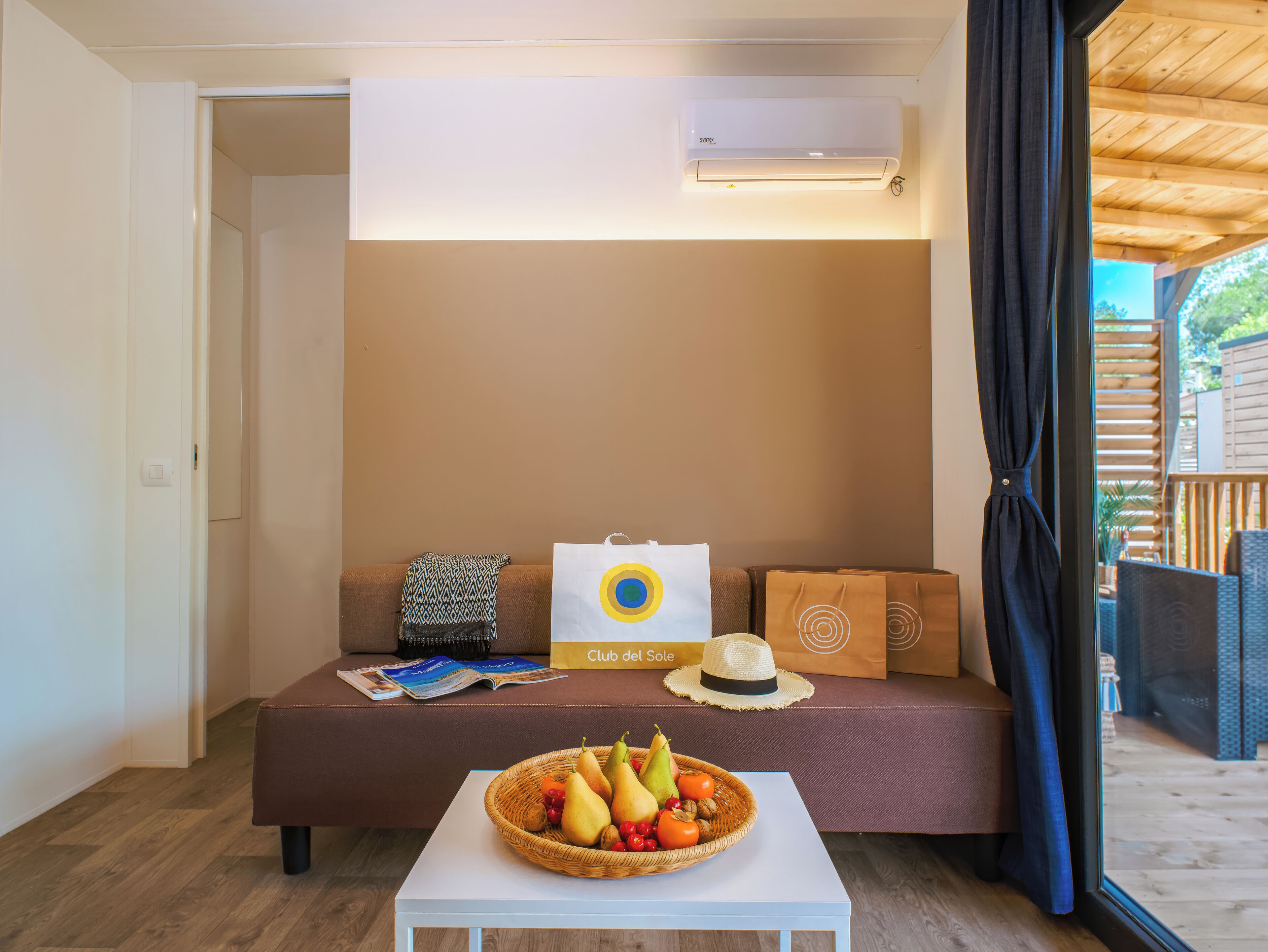 Une pièce avec un canapé marron, une table blanche sur laquelle se trouve un panier de fruits, et une fenêtre donnant sur une terrasse en bois.