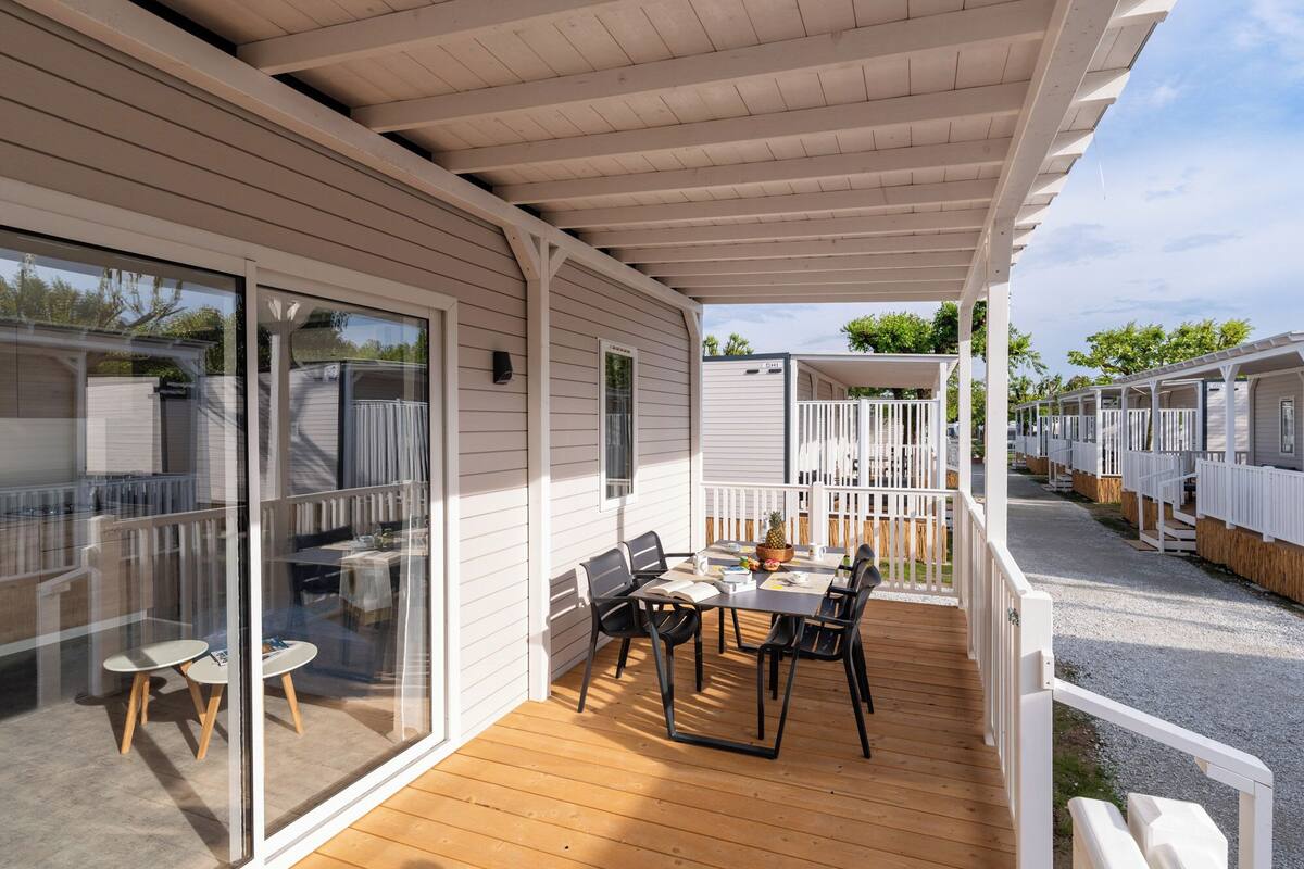 A covered outdoor area with a table and chairs, a wooden deck, and a view of adjacent mobile homes.