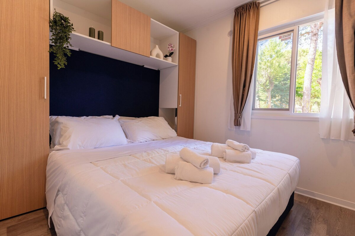 A neatly made bed with white linens and towels, a dark headboard, and a window with curtains.
