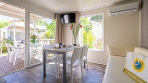 A room with a dining table, chairs, a sofa, a TV, and a window with a view of greenery. - Club Del Sole Adriatico Cervia Easy Camping Village (Cervia)
