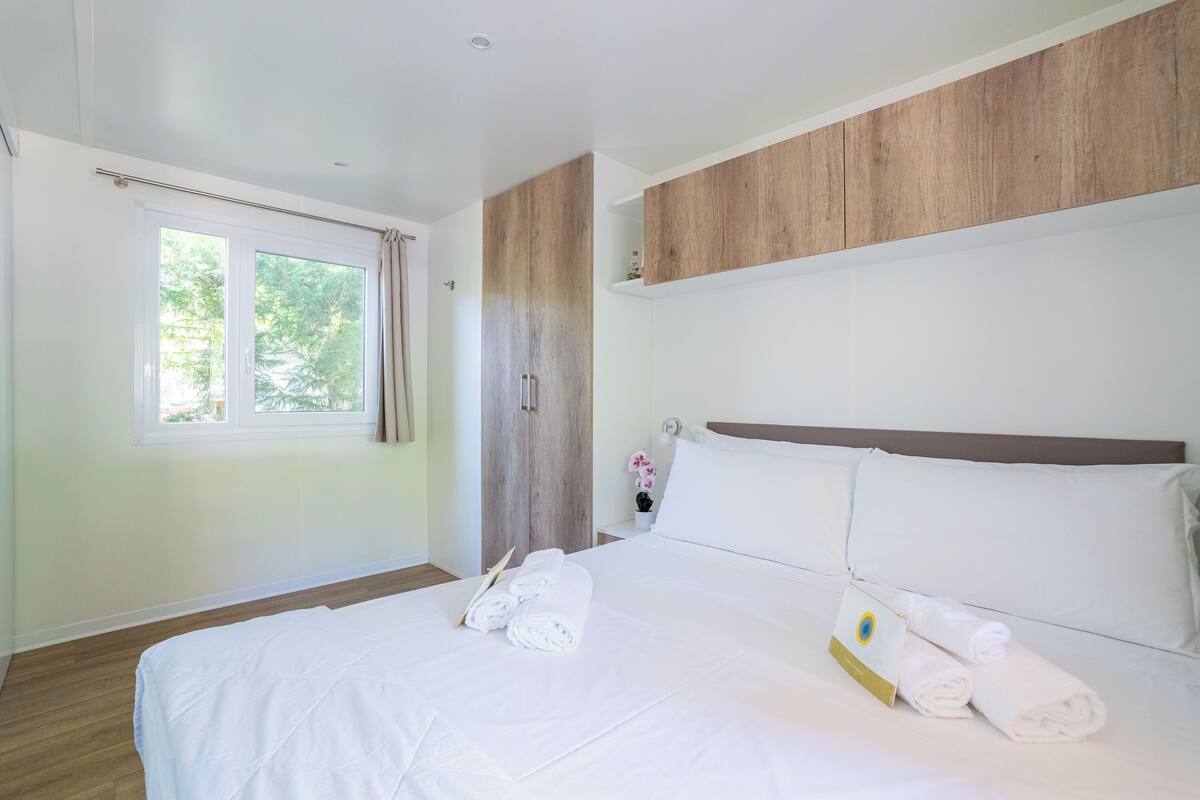 A hotel room with a large bed, a wooden wardrobe, a window with curtains, and a nightstand with a flower vase.