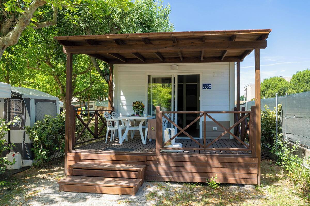 A small wooden structure with a porch, a table, and chairs, surrounded by greenery.