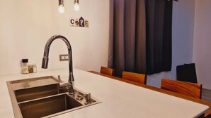 A modern kitchen with a stainless steel sink, a faucet, and a countertop. There are wooden chairs and a window with curtains.
