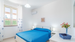 A bedroom with a bed, a nightstand, a lamp, a wall-mounted air conditioner, a window with curtains, and a vase with flowers. - Villa 'Con Tre Camere Da Letto' with Private Terrace, Shared Garden and Air Conditioning (Pachino)