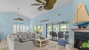 A living room with a sofa, coffee table, and patterned rug. There is a fireplace, a sailboat decoration, and a sign that reads "BEACH-HOUSE".