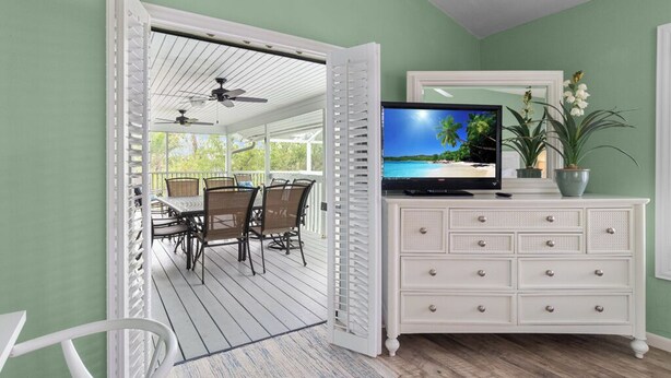 A room with a TV on a dresser, a dining area with chairs and a table, and a view of a beach through the open door.