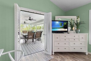 Une pièce avec un téléviseur posé sur une commode, un coin repas avec des chaises et une table, et une vue sur la plage par la porte ouverte.