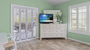 A room with a white dresser, a television on top, and a view through a window with white shutters.