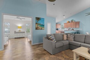 A spacious living area with a gray sectional sofa, a wooden coffee table, and a patterned rug. A kitchen with pink cabinets and a dining area with a white table are visible in the background. - Aside The Sea | Pet Friendly, Steps to the beach, 3 bedroom 2 bath with pool! (Sanibel)