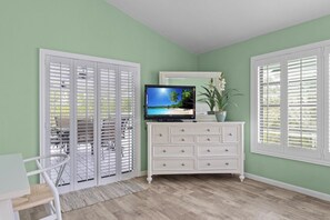 Une pièce avec une commode blanche, un téléviseur posé dessus, et une vue par une fenêtre dotée de volets blancs.