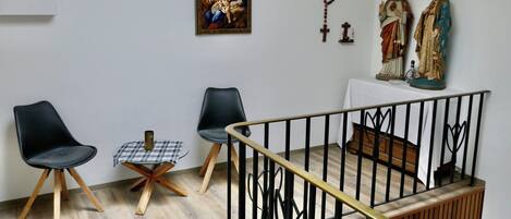 A room with a wooden floor, two black chairs, a small table with a checkered tablecloth, a wooden bench, and a railing.