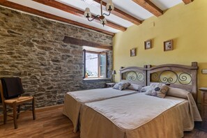 A room with two beds, stone walls, wooden beams, a window, and a chandelier.