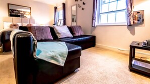 A modern living room with a black leather sofa, a small table, and a large window with curtains. - 7 Domus House Studio Apartment -  a studio that sleeps 2 guests  in 1 bedroom (Chester)