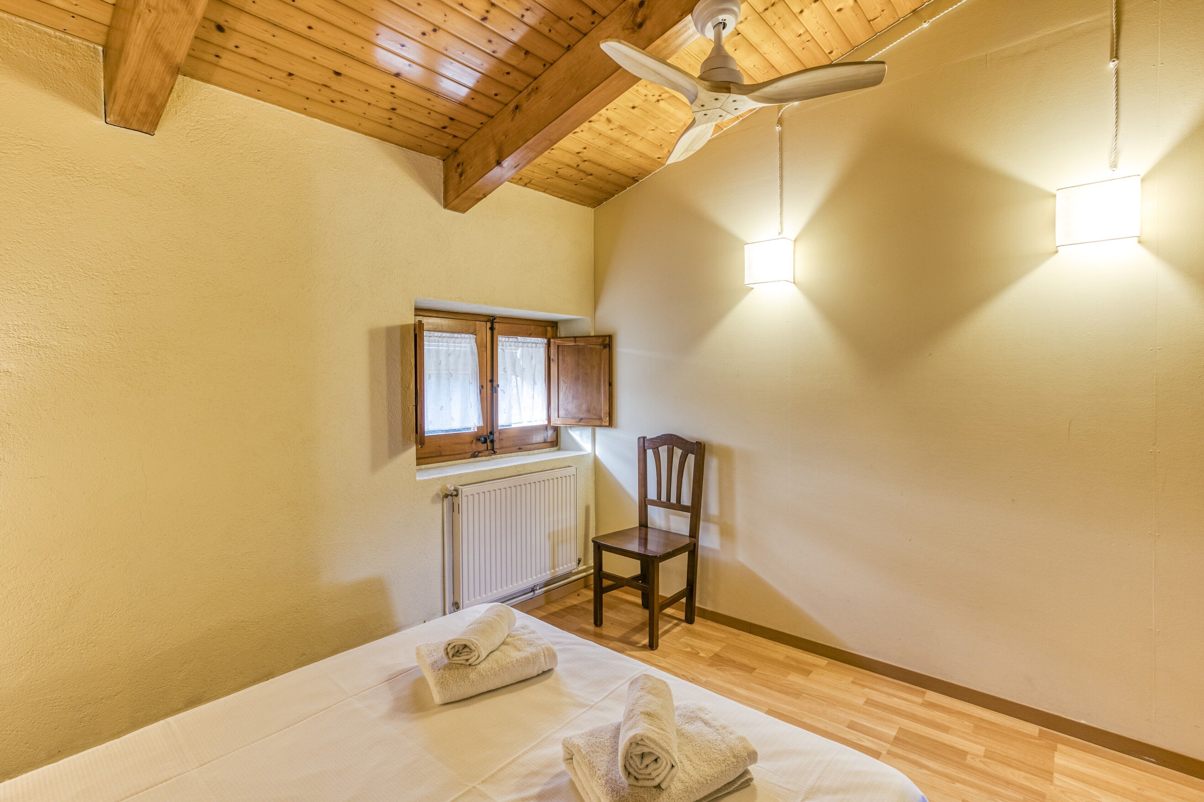 Een slaapkamer met een houten plafond, een plafondventilator, een raam met gordijnen, een radiator, een stoel en netjes opgevouwen handdoeken op het bed.
