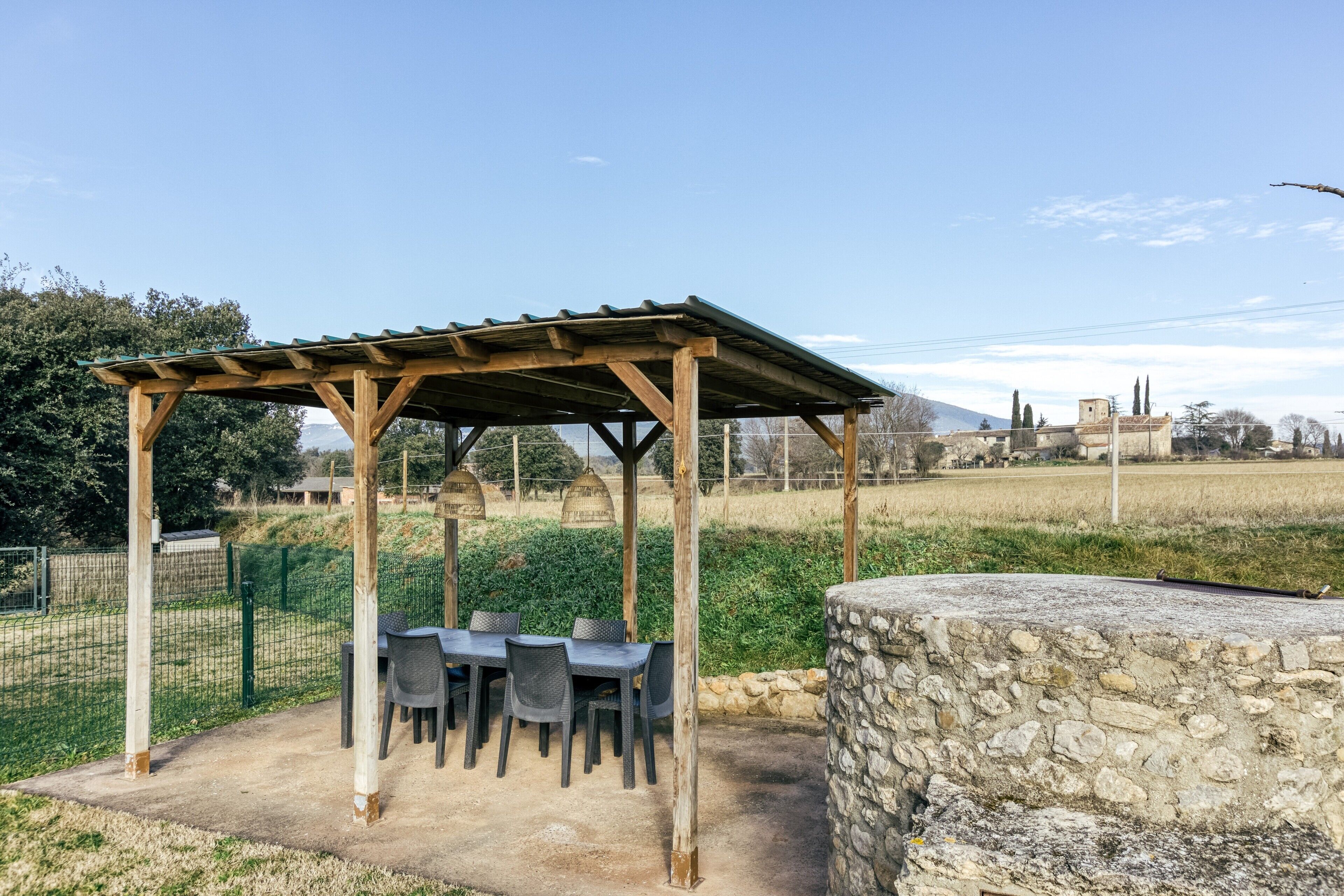 Una pérgola de madera con mesas y sillas, un muro de piedra y un área cercada con vistas a un campo y edificios a lo lejos.