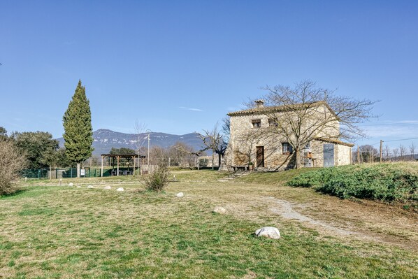 Ein Steinhaus mit Garten, ein hoher Baum und Berge im Hintergrund.