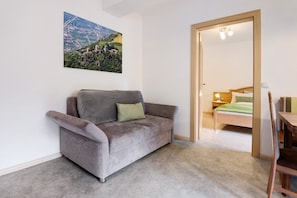 Een grijze bank met een kussen, een houten deur die leidt naar een slaapkamer met een bed en nachtkastje, en een aan de muur hangend schilderij van een landschap.