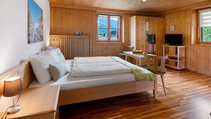 Une pièce aux murs lambrés, comprenant un lit, un bureau, une chaise, un téléviseur et une fenêtre donnant sur les montagnes.
