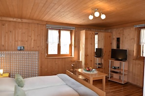 A wooden cabin room with a bed, a table with a fruit bowl, a TV on a stand, and a window with curtains.