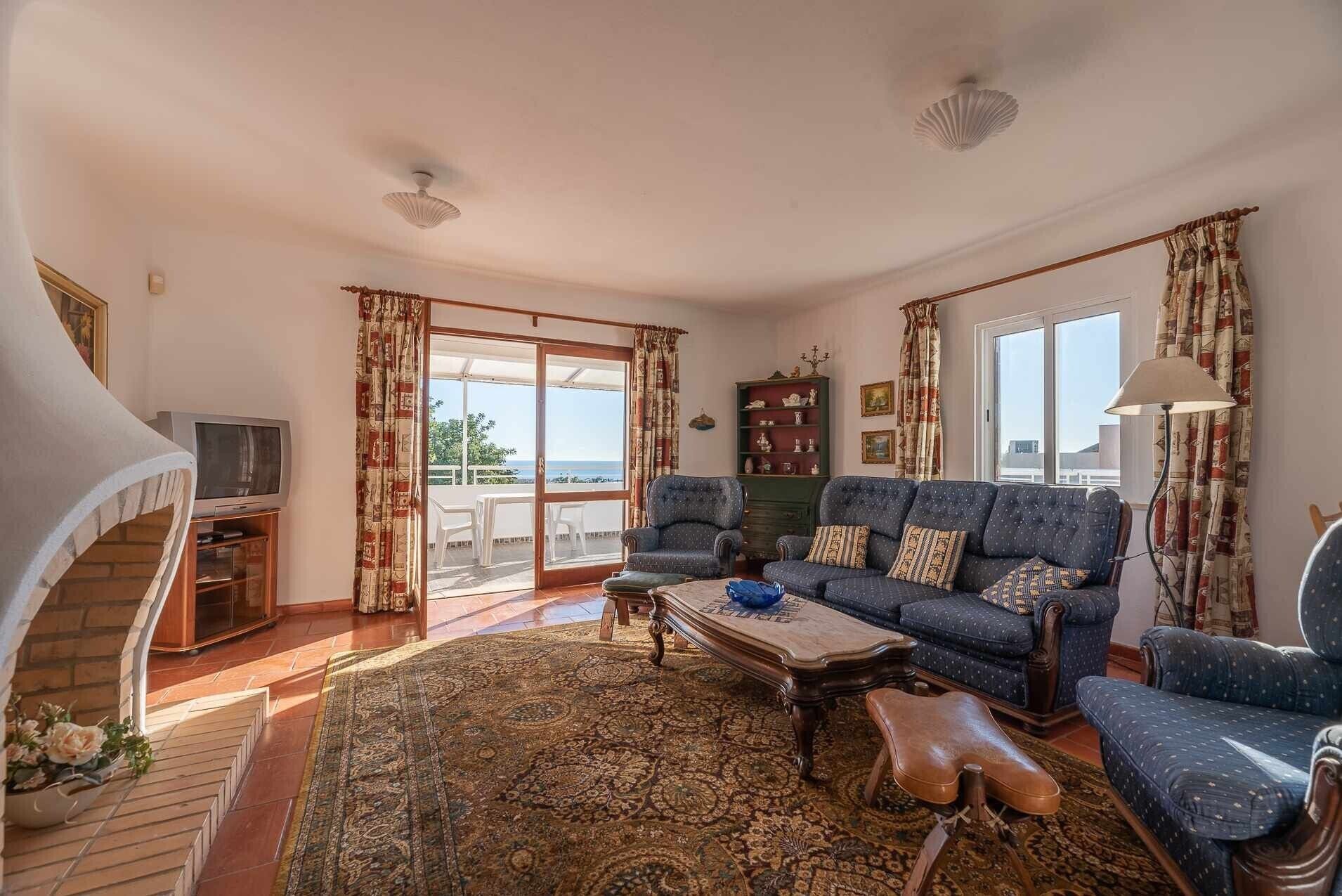 A living room with a sofa, chairs, a coffee table, a TV, and a view of the sea through a sliding door.
