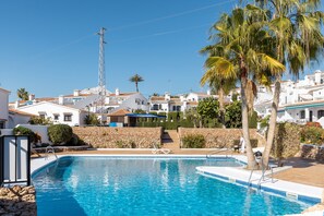 A swimming pool with a stone wall and palm trees, surrounded by white buildings with red-tiled roofs.