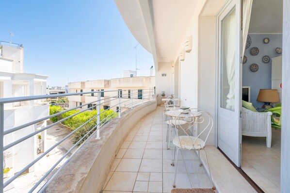 Een balkon met een witte reling, een kleine tafel met stoelen, en een rieten stoel in een kamer met een behang.