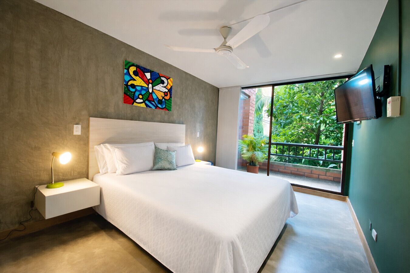 A modern bedroom with a large bed, a ceiling fan, a TV mounted on the wall, and a balcony with greenery visible through the window.