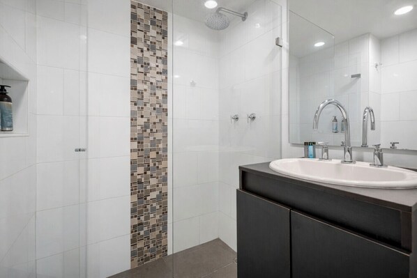 A modern bathroom with a white tiled shower, a mosaic tile accent wall, a large mirror, and a dark-toned vanity.