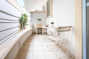 Un balcon étroit avec une balustrade blanche, une petite table avec une chaise, une plante en pot et une étagère fixée au mur, ornée d’objets décoratifs.