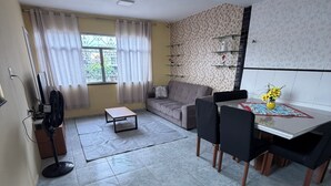 Een moderne woonkamer met een bank, eettafel en stoelen. Er staat een ventilator, een klein tafeltje en een decoratieve vaas met bloemen.