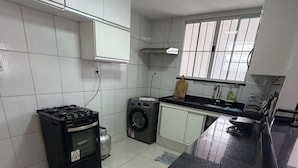Een keuken met witte kastjes, een gasfornuis, een wasmachine en een raam met jaloezieën.