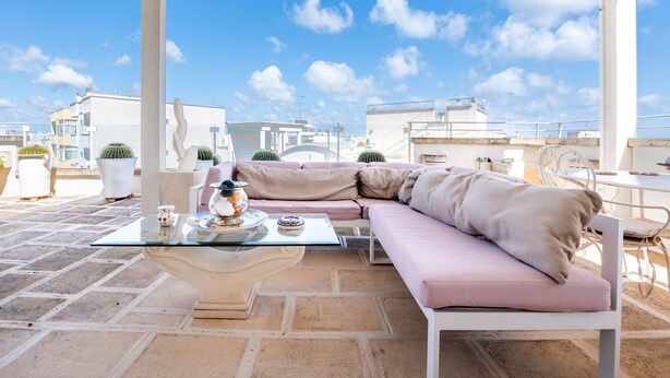 Terraço no topo do prédio com pérgola branca, sofá bege, mesa de vidro e objetos decorativos, além de cactos em vasos.