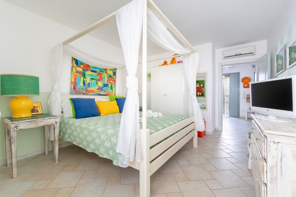 A bedroom with a four-poster bed, a television, a bedside table with a lamp, and a colorful abstract painting on the wall.