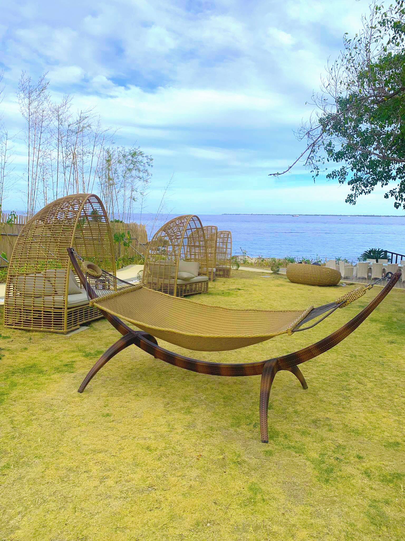A hammock chair in a grassy outdoor area with wicker furniture and a view of the sea.