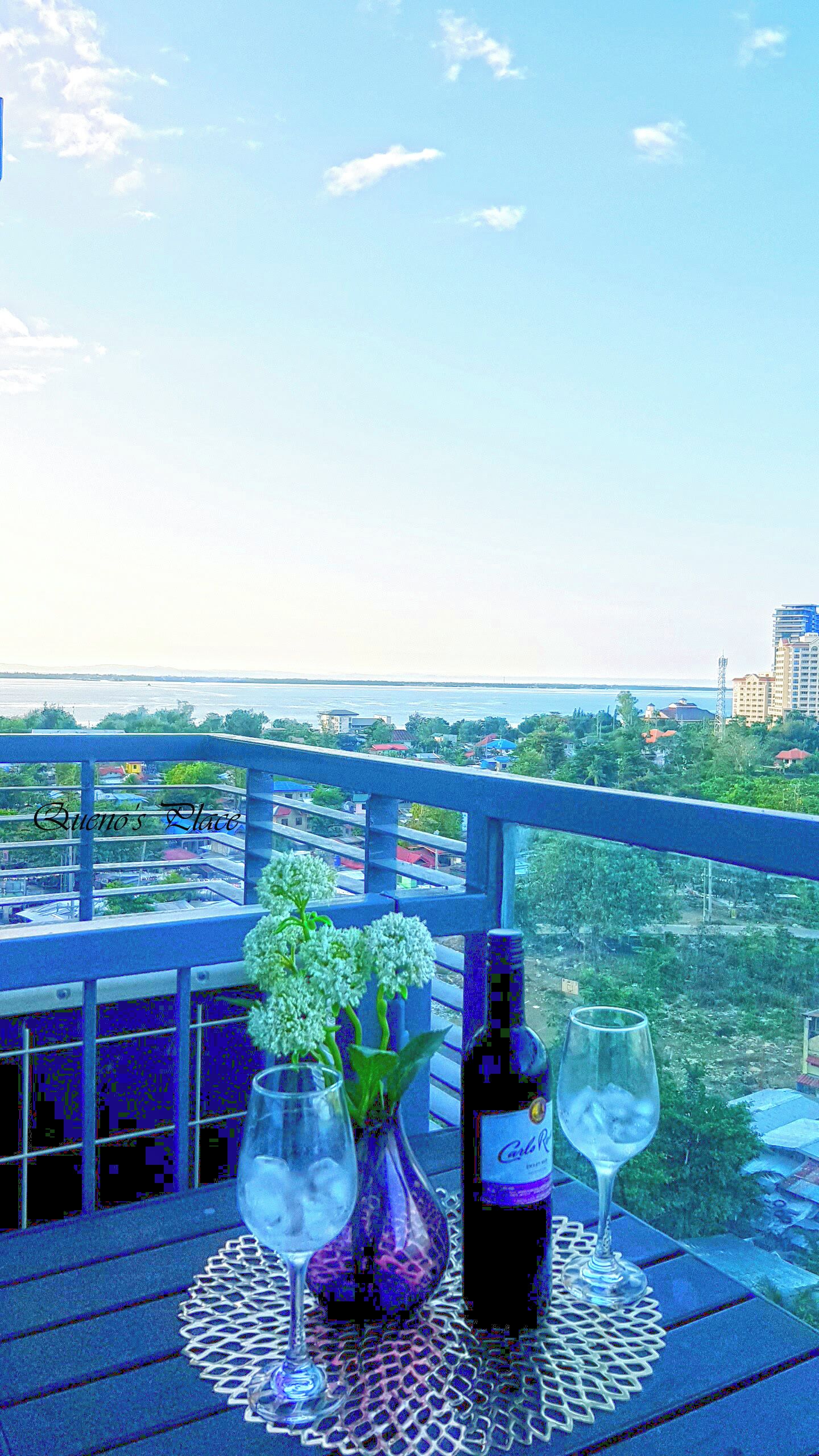 En balkon med et bord dækket til to personer, komplet med en vinflaske og to glas, med udsigt over et bybillede med bygninger og et vandareal.