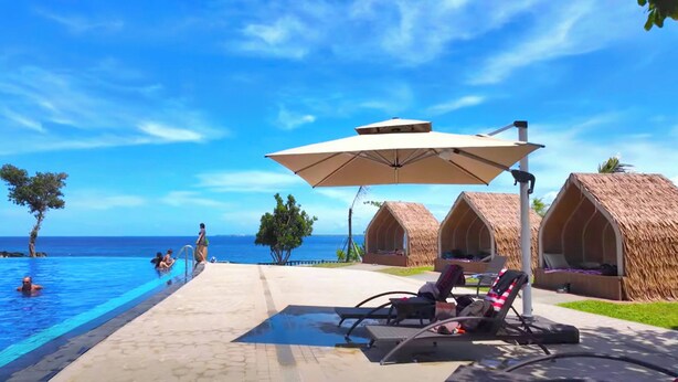 A poolside area with lounge chairs and umbrellas, a clear blue sky, and thatched-roof huts in the background. - One Pacific Residence 1BR Ocean View (Lapu-Lapu)