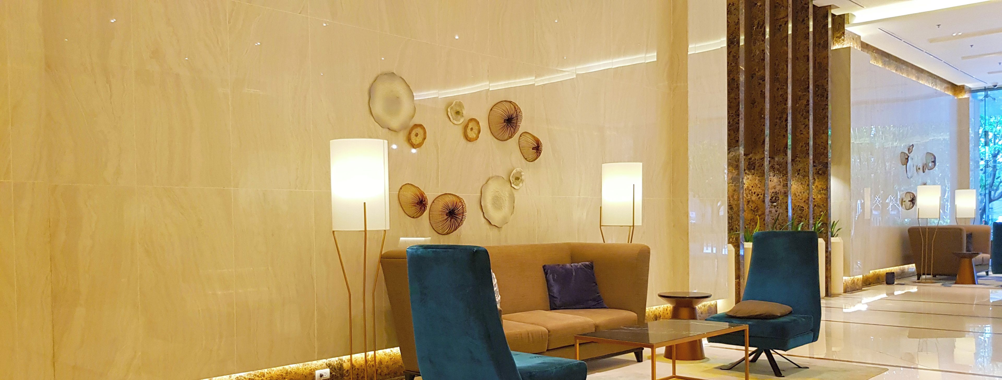 A modern lobby with a beige marble wall, a beige sofa, two blue armchairs, a glass coffee table, and a floor lamp.