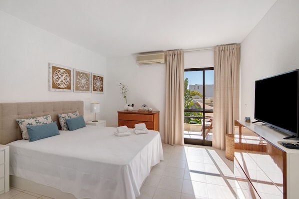 A hotel room with a bed, a TV, a desk, and a balcony with curtains.