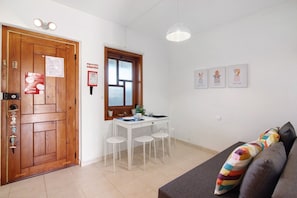 A room with a wooden door, a dining table with chairs, a window, a sofa, and wall-mounted pictures.