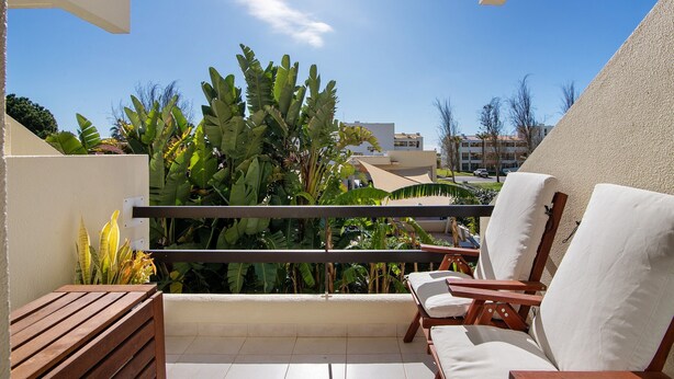 A balcony with a wooden bench, two white cushioned chairs, and a view of greenery and buildings. - 'Golden' Apartment with Shared Pool, Wi-Fi and Air Conditioning (Cabanas de Tavira)