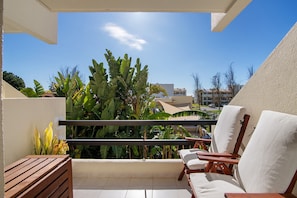 A balcony with a wooden bench, two white cushioned chairs, and a view of greenery and buildings.